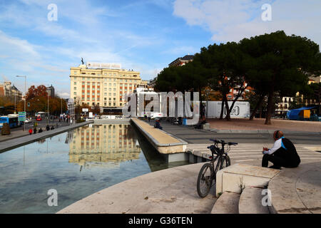 Mann, der in Jardines del Descubrimiento, Plaza de Colon, Madrid, Spanien Kopfhörer neben dem Fahrrad trägt. Hotel Fénix Gran Meliá im Hintergrund. Stockfoto