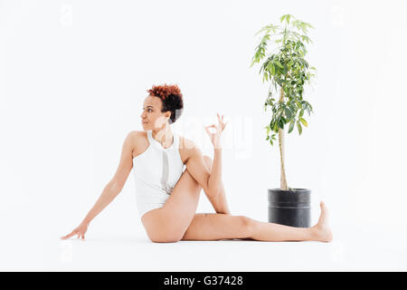 Ziemlich junge Afroamerikanerfrau entspannen und praktizieren von Yoga in der Nähe kleiner Baum im Topf auf weißem Hintergrund Stockfoto