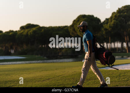 schöne orientalische Golfer Tragetasche und zu Fuß zum nächsten Loch am Golfplatz auf wunderschönen Sonnenuntergang im Hintergrund Stockfoto