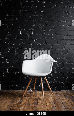 Weißer Stuhl stehend im Raum auf braunem Holzboden über schwarze Ziegelmauer Stockfoto