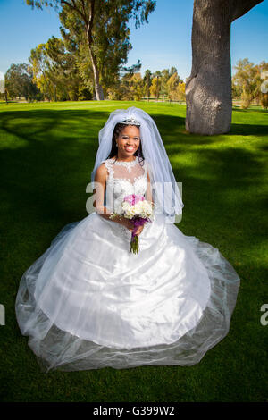 Eine schöne, African American Braut kniet vor ihrer Hochzeit für ein Porträt.  Sie ist umgeben von üppigen, grünen Rasen. Stockfoto