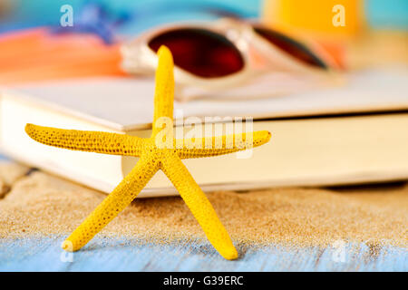 Nahaufnahme von einer gelben Seestern auf ein Haufen Sand, und eine Sonnenbrille an einem Buch auf einer rustikalen blau Holzfläche gelegt Stockfoto