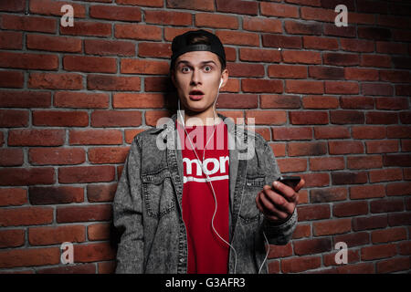 Überrascht Jugendlicher Blick in die Kamera und Musikhören gegen Mauer Stockfoto