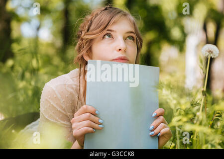 Niedliche nachdenkliche junge Frau auf dem Rasen liegen und holding blank Magazin Stockfoto