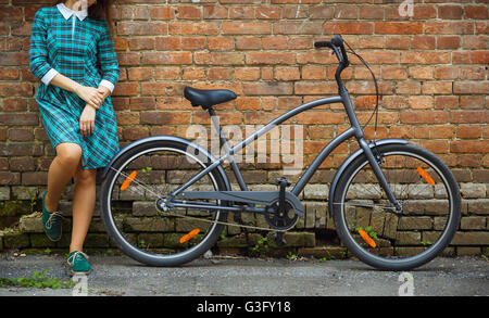 Schwarze Retro-Fahrrad und ein Mädchen in ein grünes Kleid in der Nähe von es auf dem alten Ziegel Wand Hintergrund Stockfoto