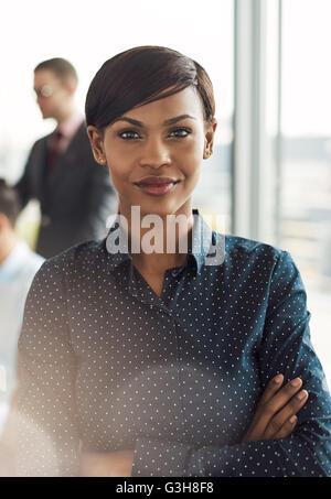 Schöne junge grinsende Business-Frau im Büro mit Polka Dot Bluse, verschränkten Armen und zuversichtlich Ausdruck als starkes Licht fl Stockfoto