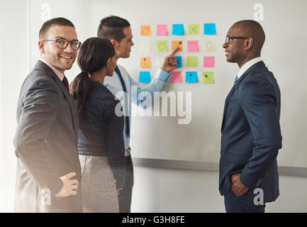 Gruppe von Geschäftsleuten, die Lösung eines Rätsels auf bunte Notizzettel an einer Wand mit einem junge männliche Führungskraft auf der Rückseite mit Blick auf smi Stockfoto