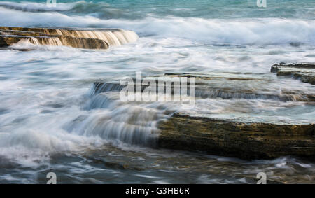 Starke Wellen, die mit der Kraft der Meer rock Platten erstellen, kleinen Wasserfällen und glatte Ströme von Wasser. Stockfoto