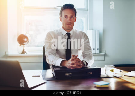 Executive Business-Mann an seinem Schreibtisch mit Autorität in die Kamera schaut die Hände gefaltet auf Aktentasche am Schreibtisch Stockfoto