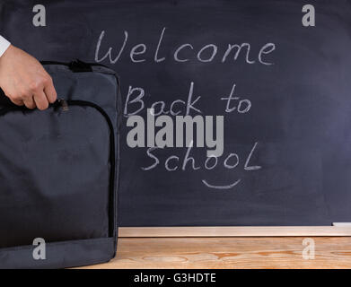 Hand mit Rucksack auf Desktop. Gelöschten schwarzen Tafel im Hintergrund zusammen mit Willkommens-Nachricht. Stockfoto