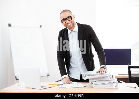 Junge hübsche lächelnd Geschäftsmann stehend auf dem Schreibtisch im Büro Stockfoto