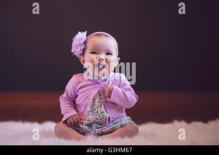 Babymädchen sitzen auf Teppich tragen Blütenkopf Band Blick auf Kamera Mund öffnen Sie lächelnd Stockfoto