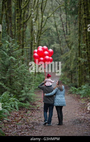 Rückansicht der Eltern mit Kleinkind Tochter auf Schultern und Haufen rote Luftballons im Wald spazieren Stockfoto