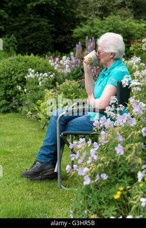 Eine reife Frau sitzt in einem Gartenstuhl, trinken aus einem Becher Tee, umgeben von Sommerblumen. Stockfoto