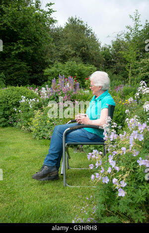 Eine reife Frau sitzt in einem Landschaftsgarten voller Blumenrabatten umgeben. Sie sitzt mit einer Tasse Tee entspannen. Stockfoto