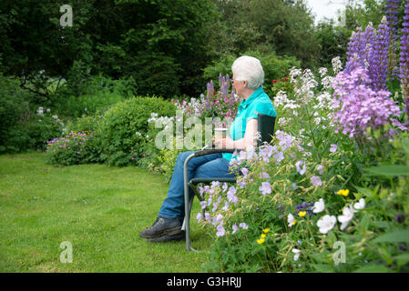 Eine reife Frau sitzt in einem Landschaftsgarten voller Blumenrabatten umgeben. Sie sitzt mit einer Tasse Tee entspannen. Stockfoto