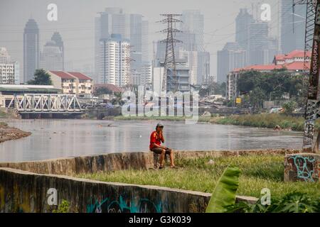 Jakarta, Indonesien. 18. April 2016. Ein Mann raucht am Ufer des Flusses, wie Hochhäuser auf dem Hintergrund gesehen werden. © Garry Andrew Lotulung/Pacific Press/Alamy Live-Nachrichten Stockfoto