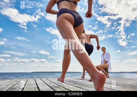 Junge Frau sitzt auf Post auf hölzernen Pier, gerade Freunde laufen in Richtung Rand, niedrigen Winkel Ansicht Stockfoto