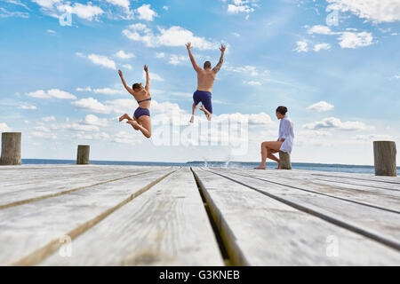 Junge Frau sitzt auf Post auf hölzernen Pier, Freunde beobachten, wie sie ins Meer springen Stockfoto