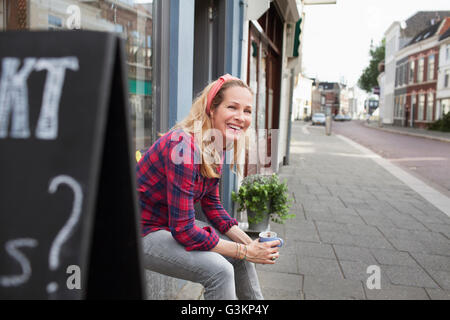 Frau sitzt auf Shop Fensterbank halten Kaffee Tasse aussehende Weg Lächeln Stockfoto