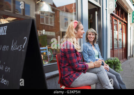 Frauen sitzen auf Stühlen vor Geschäft hält lächelnd Kaffeetassen Stockfoto