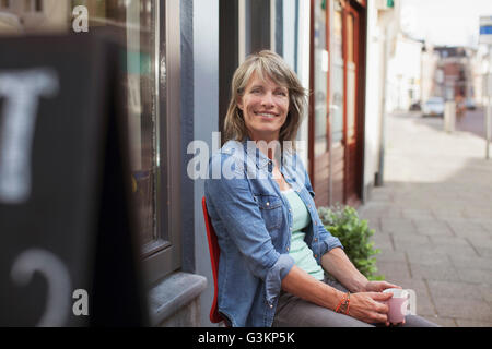 Frau sitzt auf dem Stuhl vor Geschäft halten Kaffeetasse Blick auf die Kamera zu Lächeln Stockfoto