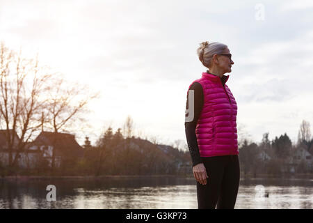 Frau trägt rosa Thermoweste vor Teich wegschauen Stockfoto