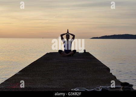 Junge Frau sitzt am Pier in Yogaposition, in der Dämmerung Stockfoto