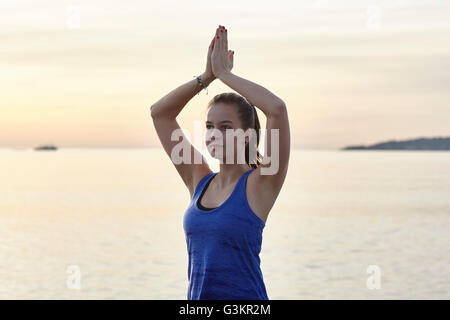 Junge Frau neben Meer in Yogaposition bei Sonnenuntergang Stockfoto