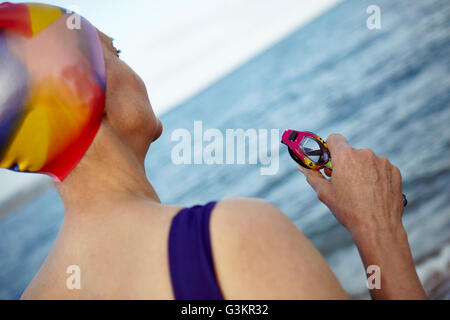 Reife Frau trägt Badehose, Schwimmen Hut und halten schwimmen Schutzbrillen, stehend am Meer, Rückansicht Stockfoto