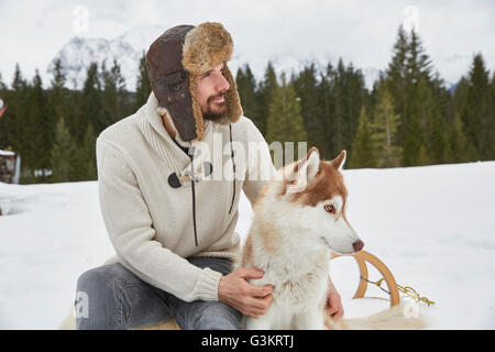 Junger Mann tragen Trapper Hut Petting husky im Schnee, Elmau, Bayern, Deutschland Stockfoto