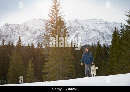 Junger Mann zu Fuß bergauf mit Husky in der tief verschneiten Landschaft, Elmau, Bayern, Deutschland Stockfoto