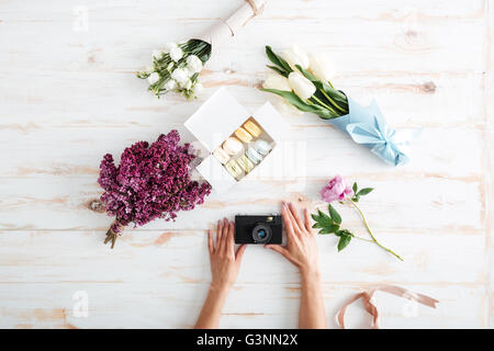 Hände der jungen Frau mit Fotokamera auf Holztisch mit französische Macarons und Blumen Stockfoto