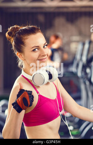 Glückliche Mädchen mit Kopfhörern auf Cardio-Training im Fitness-Center. Lächelndes schöne Mädchen zeigt Daumen oben im Cardio-Bereich Stockfoto