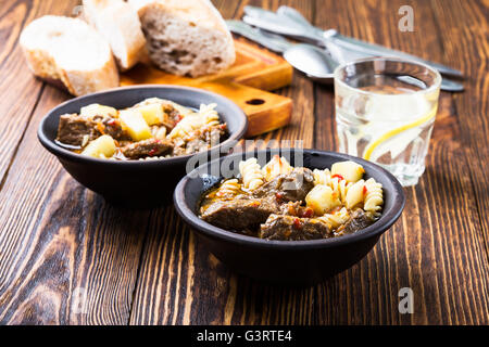 Rindergulasch mit Nudeln und Kartoffeln in Keramikschale auf Holztisch Stockfoto