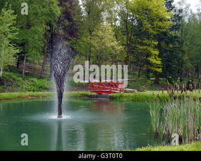 Skulptur, Brunnen und rote Brücke in Green Lake an der Himalaya Garten & Skulpturenpark, North Yorkshire, England UK. Stockfoto