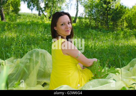 Das Mädchen sitzt auf einer Wiese im Sommer Holz eine Pause, das Mädchen, das Holz, Sommer, einen Rasen, gelb, ein Kleid, Grün hat, gelb Stockfoto