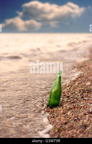 Einzigen leeren grünen Glas Flasche aufrecht stehend an einem steinigen Strand mit blauem Himmel in der Ferne. Fläche für Text zu kopieren und Stockfoto