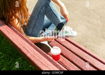 Junge Frau mit roten Einweg-Pappbecher Kaffee mit TabletPC sitzen auf einer roten Bank in einem park Stockfoto