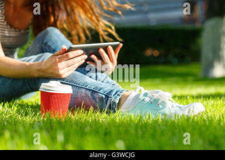 Junge Frau mit roten Einweg-Pappbecher Kaffee mit Tablet-pc auf einem grünen Rasen in einem park Stockfoto