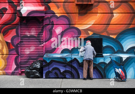 Ältere asiatische Frau sammeln Dosen und Plastikflaschen aus Mülltonnen vor Streetart Wandbild in Nolita in New York City Stockfoto