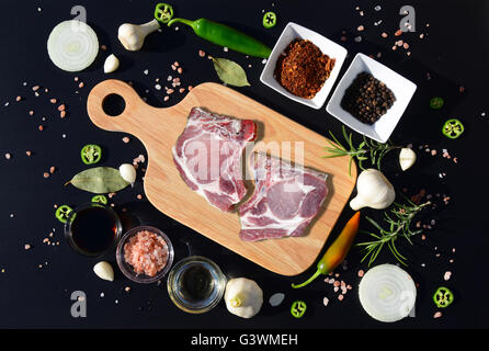 Rohes Schweinefleisch auf einer Cutting Board und Pfeffer, Lorbeerblatt, Rosmarin, Zwiebeln, Salz, Olivenöl, Soße auf schwarzem Hintergrund Stockfoto