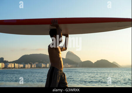 RIO DE JANEIRO - 5. April 2016: Ein junger brasilianischen Mann am Strand der Copacabana gleicht einem Surfbrett auf dem Kopf bei Sonnenaufgang. Stockfoto