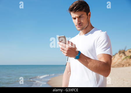 Hübscher Junge Sportler stehend und mit Handy am Strand Stockfoto