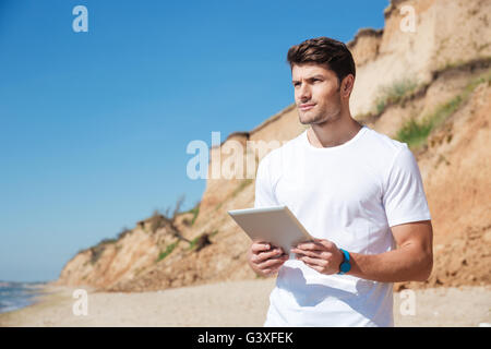 Nachdenklicher junger Mann stehend und mit Tablet am Strand Stockfoto