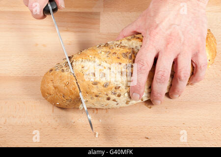 Mans Hände schneiden zu einem Honig beschichtet Sonnenblumen ausgesät Laib frisches Brot auf ein Schneidbrett aus Holz Stockfoto