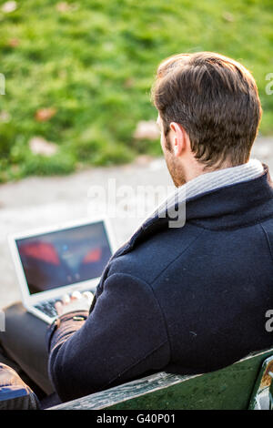 Schöne elegante Geschäftsmann sitzt auf einer Holzbank, arbeiten im Freien in einem städtischen Park, die Eingabe von Informationen auf seinem Laptop comp Stockfoto