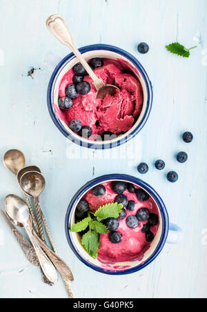 Hausgemachte Heidelbeer-Eis schöpft mit frischen Beeren und Minze in Emaille Becher über hellblauen Hintergrund, Draufsicht, sele Stockfoto