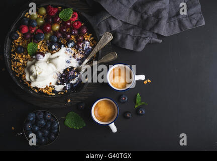 Gesundes Frühstück. Hafer-Müsli Crumble mit frischen Beeren, Samen und Eis in Bratpfanne Eisenpfanne auf dunklen Holzbrett und Tassen Stockfoto