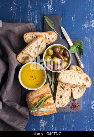 Mediterrane snacks Satz. Oliven, Öl, Kräuter und Ciabatta Schnittbrot auf schwarzem Schiefer Steinplatte über bemalte dunkel blaue Hinterg Stockfoto